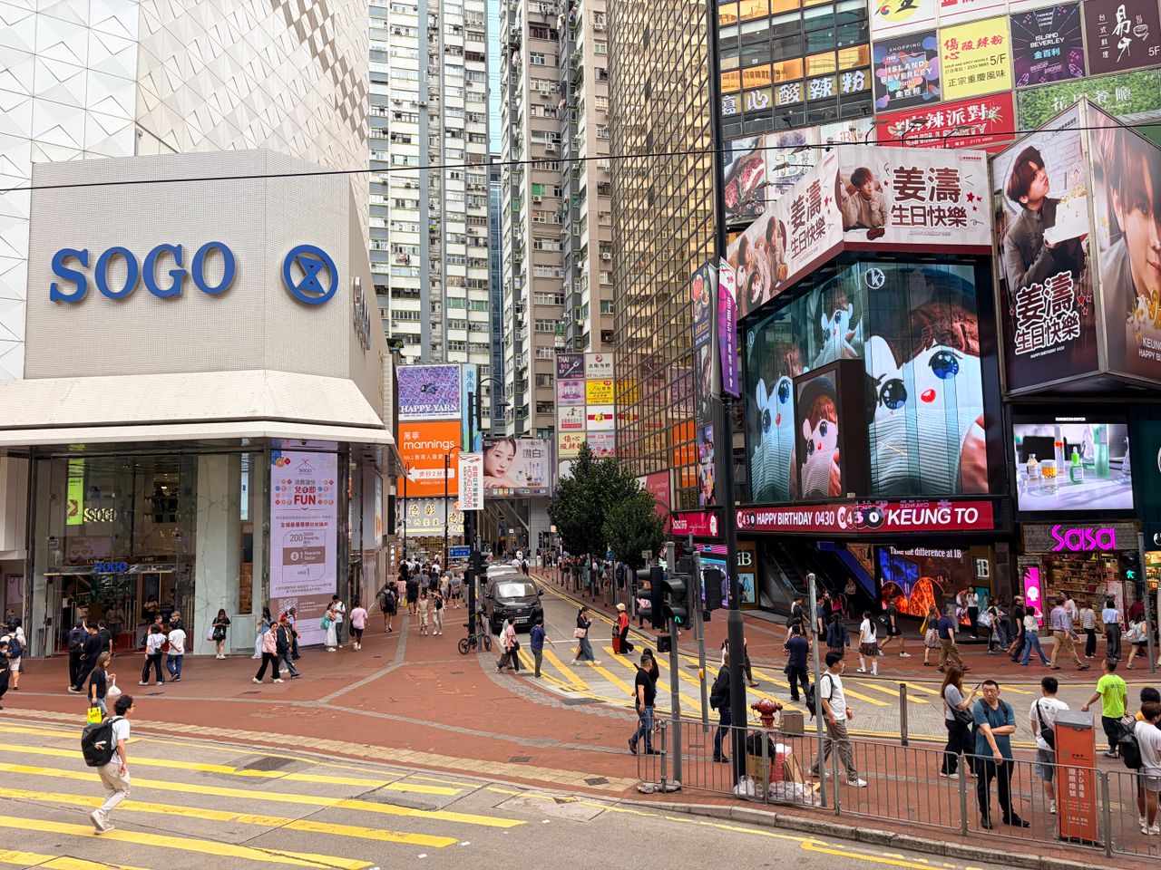 gallery-4 Vibrant city scene depicting a busy street near Sogo Department Store in Hong Kong, with digital billboards and pedestrians.