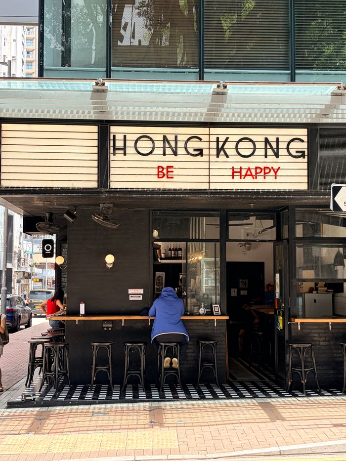 about-02 Street view of a café in Hong Kong with a 'Hong Kong Be Happy' sign, promoting positivity.