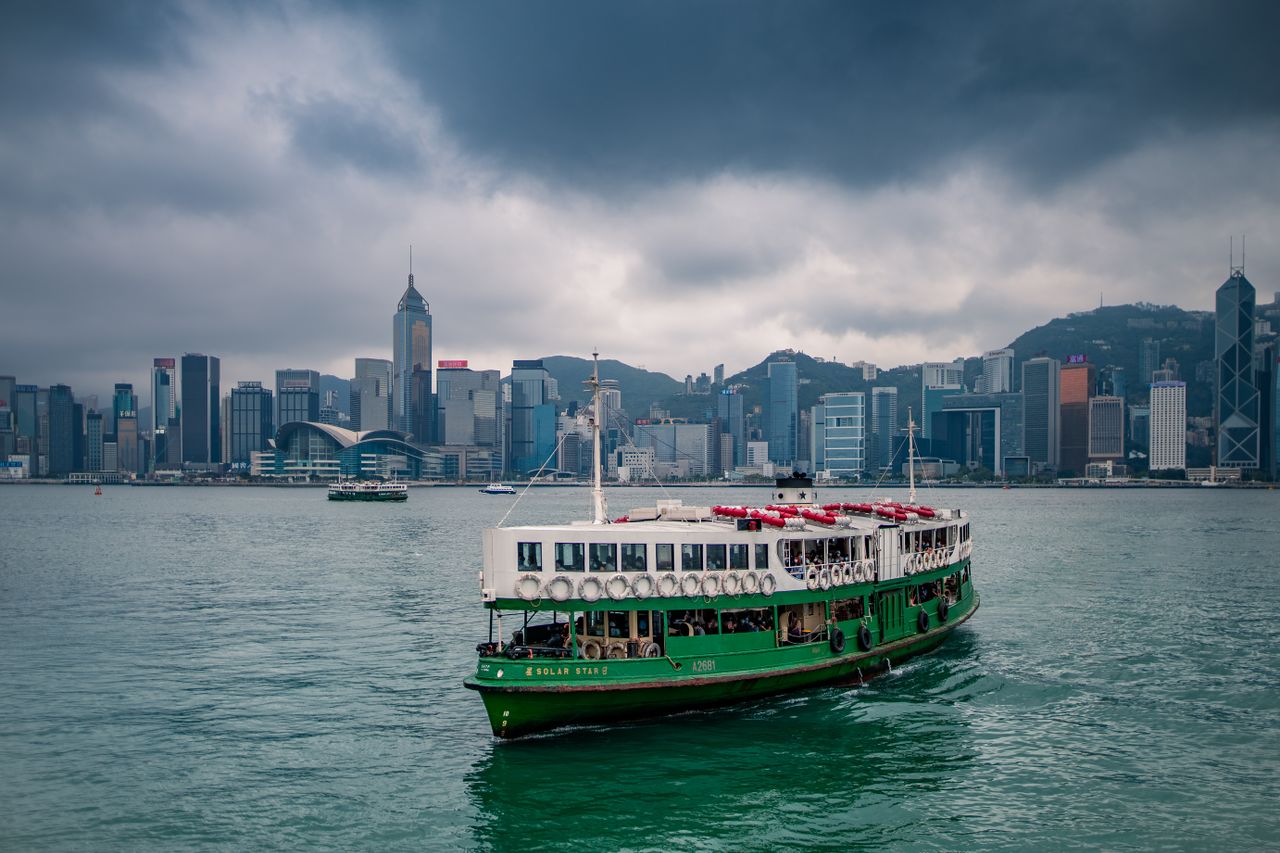 services-01 Scenic view of a traditional ferry on Victoria Harbour with Hong Kong skyline under cloudy skies.