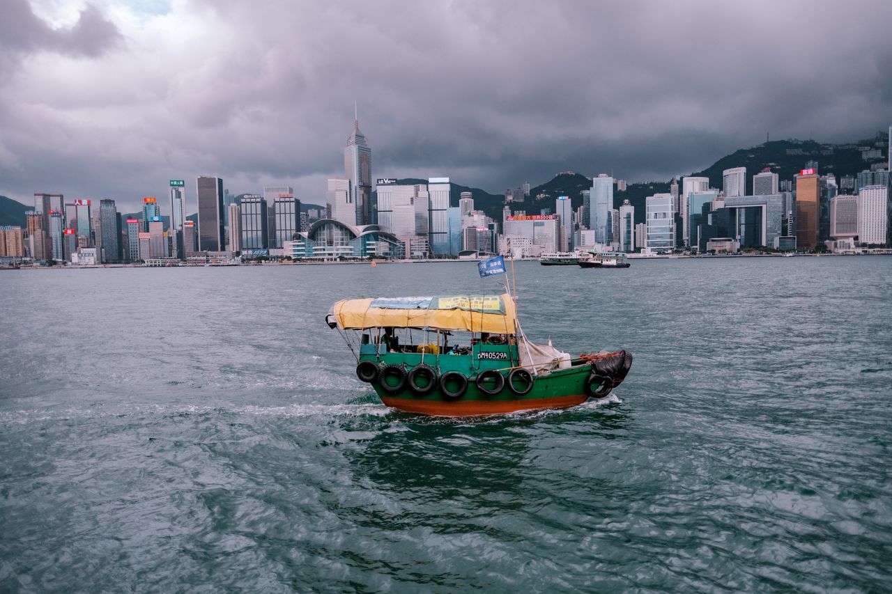 hero-img-01 A traditional boat sails in Victoria Harbour with Hong Kong's iconic skyline under dramatic clouds.